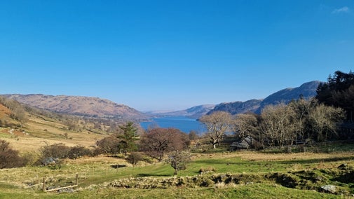 Early spring view of Ullswater Lake with surrounding fells with Seldom Seen cottages to the left on a sunny day with a blue sky.